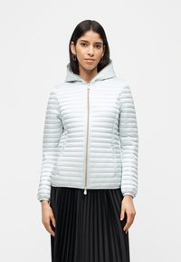 Γυναίκα που φοράει ανοιχτό γκρι μπουφάν με φερμουάρ και κουκούλα, καθώς και μαύρη πλισέ φούστα, στέκεται μπροστά σε απλό λευκό φόντο.