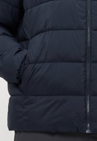 Puffet jakke i mørk marineblå med vandrette quiltede sømme, frontlynlås og sidelommer, båret med en hånd i lommen.