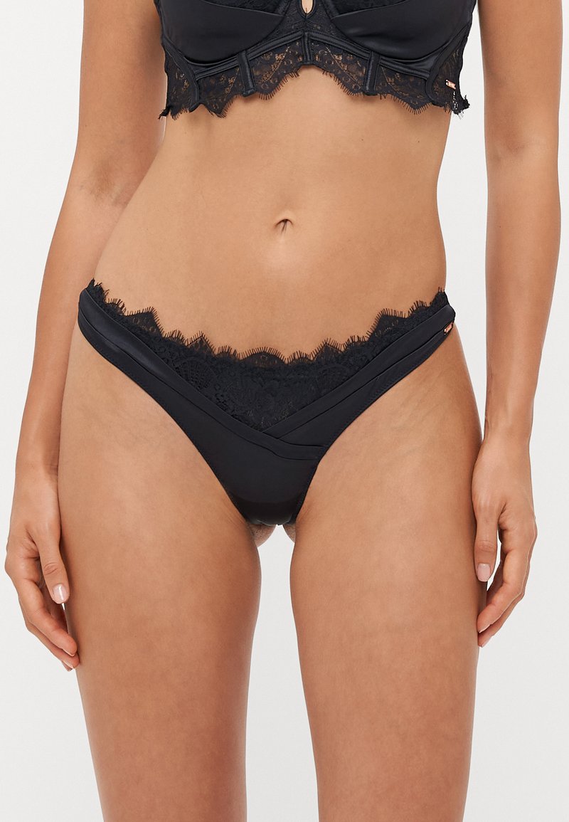 String nero realizzato in tessuto liscio, con un bordo in pizzo lungo l'orlo della vita e un sottile design incrociato sul davanti.