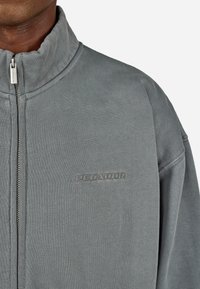 Vyras vilki pilką zip-up svarką su aukštu apykakle ir subtiliu siuvinėtų tekstu "PECADOR" kairėje krūtinės pusėje.