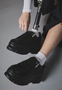 Baskets à plateforme noires avec une semelle épaisse, cuir texturé, fermeture à lacets et détail de fermeture éclair. Portées avec des chaussettes blanches et une jambe prothétique.