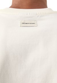 Crème kleurig t-shirt met een geribbelde textuur. Bevat een label op de achterkant met de tekst "DON'T WASTE CULTURE®" in zwarte letters.