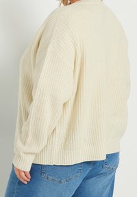 En person som bär en krämfärgad tjockstickad tröja och blå denimjeans, visad från sidan och bakifrån med en avslappnad passform.