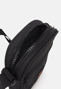 Black fabric crossbody bag featuring a zippered closure, textured surface, and an adjustable strap. Includes a front patch detail.