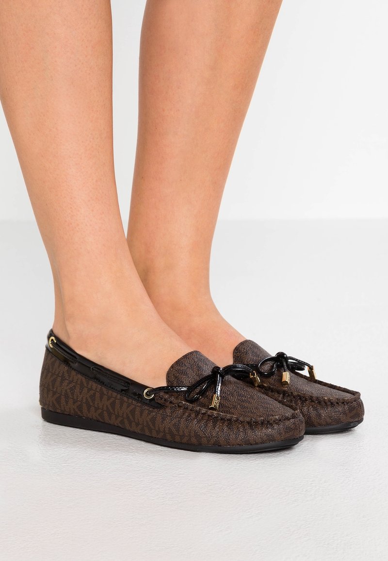 Pieds portant des mocassins marron à enfiler avec des accents de nœuds noirs et des embouts dorés, présentant un motif de logo répétitif subtil sur un fond clair.