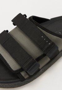 Melnas un pelēkas čības ar teksturētām siksnām, diviem regulējamiem Velcro aizdariem un mīkstinātu zoli ar smalku rakstu.
