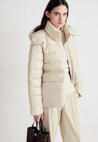 Casaco puffer bege com gola alta e capuz incorporado, apresentando fecho com zíper, ligeiro brilho e detalhes em botão. Usado sobre uma blusa crème.