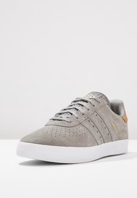 Grå mockaskor med perforerat mönster, vit gummisul, tre randig design och en brun läderhælaccent.