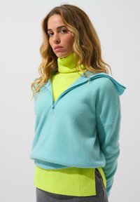 Heller blauer Strickpullover mit einer Kapuze mit Reißverschluss, �über einem leuchtend gelben Rollkragenpulli getragen. Strukturiertes Material und kontrastierende Farbbänder an den Bündchen.