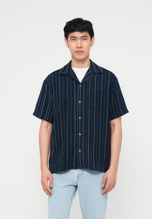 Junger Mann trägt ein marineblaues, kurzärmeliges Hemd mit weißen vertikalen Streifen und hellblaue Jeans, steht vor einem einfarbigen Hintergrund.