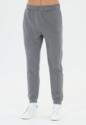 Homme portant un pantalon de jogging gris et des baskets blanches debout devant un fond blanc uni.