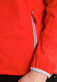 Chaqueta de softshell roja con un bolsillo con cremallera, puños elásticos y acentos grises en los puños. La tela parece suave y ligeramente elástica.