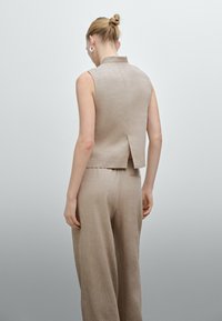 Beige linne ärmlös topp med hög krage och slits bak, kombinerad med lösa beige byxor. Slät textur, enkel design.