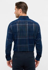 Camicia a quadri blu navy con bottoni, caratterizzata da una texture morbida, maniche lunghe e colletto. La vista posteriore mostra un mix di quadrati blu, verdi e marroni.
