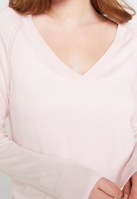 Světle růžový svetr se zkříženým výstřihem a dlouhými rukávy, s vroubkovanými manžetami a jemnou strukturou. Tkanina se zdá být měkká a přiléhavá, vhodná na vrstvení.