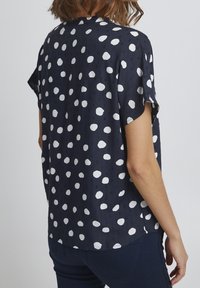 Short-sleeve blouse in navy with white polka dots. Lightweight, smooth fabric, rounded hem, casual fit with side slits for comfort.