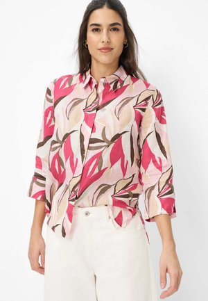 Frau trägt ein langärmeliges Hemd mit Knopfleiste, bedruckt mit Muster aus pinken, braunen und beigen Blättern, kombiniert mit hoch taillierten weißen Hosen.