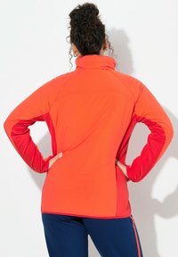 Czerwona sportowa kurtka z wysokim kołnierzem, długimi rękawami i kontrastującymi czerwonymi akcentami. Gładka faktura i dopasowany fason. Widok z tyłu.