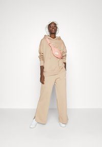 Sudadera beige con un bolsillo frontal, pantalones de pierna ancha beige a juego y una riñonera rosa con un logo blanco, complementada por zapatillas blancas.