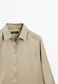 Camicia beige con colletto classico, tessuto morbido e maniche corte con un sottile design sulle spalle. Etichetta del marchio visibile.