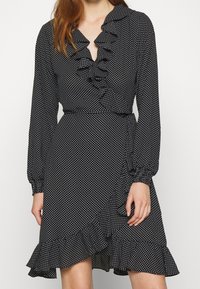 Vestido negro con lunares, de mangas largas, con escote volado y un dobladillo en capas. Hecho de una tela ligera con una cintura ajustada.