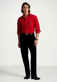 Κόκκινο πουκάμισο με κουμπιά και μακριά μανίκια, με διακοσμητικό τσεπάκι στο στήθος, συνδυασμένο με μαύρο παντελόνι και μαύρα παπούτσια.