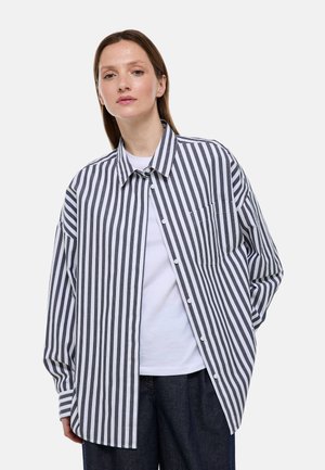 Femme portant une chemise ample à rayures verticales noires et blanches boutonnée, par-dessus un haut blanc et un pantalon foncé, debout devant un fond uni.