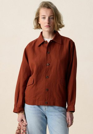 Vrouw die een roestkleurig jasje met knopen en lichtblauwe spijkerbroek draagt, met een patroon clutch in haar linkerhand tegen een effen achtergrond.