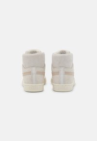 Ein Paar beige Wildleder High-Top Sneakers mit strukturierten Gummisohlen, von hinten auf einem weißen Hintergrund betrachtet.