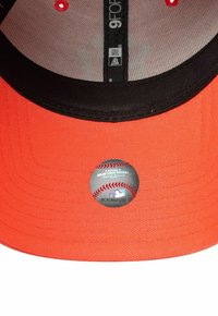 Orangene Baseballmütze mit einem schwarzen Schirm; auf der Unterseite befindet sich ein runder MLB-Aufkleber. Strukturierter Stoff mit sichtbaren Nählinien.