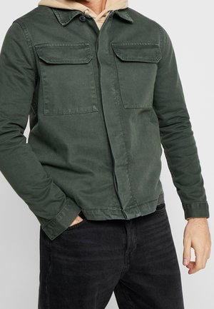 Mann trägt eine dunkelgrüne, geknöpfte Jacke mit zwei Brustklapptaschen über einem beige Hoodie und schwarzen Jeans, Hand in der Tasche.