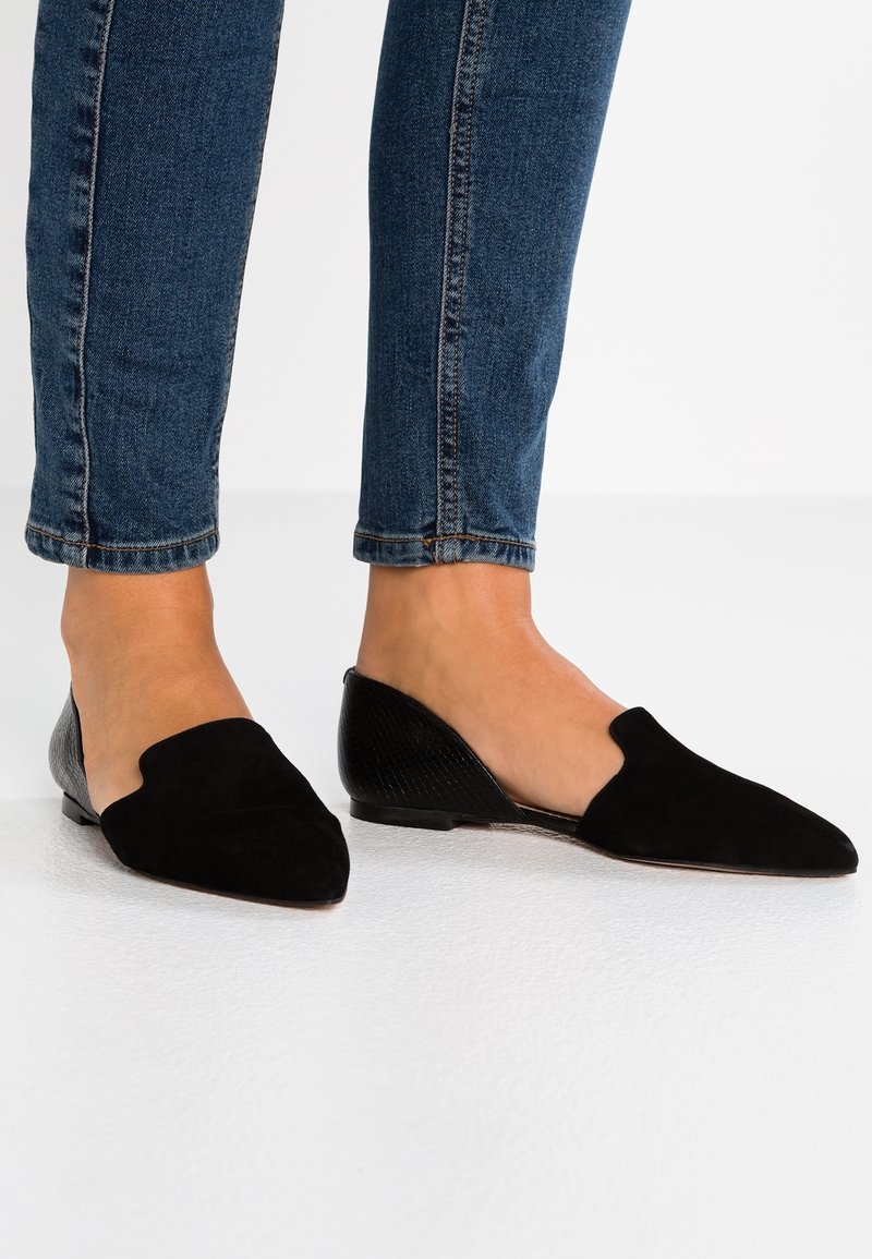 Des ballerines en suède noir avec un bout pointu, présentant un design découpé et un dos lisse en matériau texturé, associées à un jean en denim foncé.