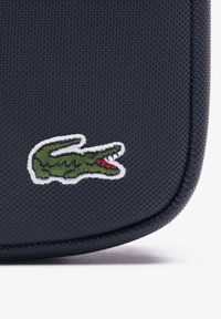 Bolso negro texturizado con un logo de cocodrilo bordado en verde y rojo sobre un contorno blanco. Esquinas redondeadas con detalle de costura.