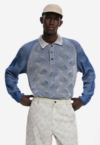 Polo à manches longues bleu et blanc avec motif à rayures ondulées, doté d'un col à boutons, assorti à un pantalon de couleur claire.