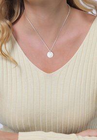 Srebrny naszyjnik z wisiorkiem o fakturowanej, okrągłej zawieszce na cienkim łańcuszku, noszony na jasnobeżowej, prążkowanej bluzce.