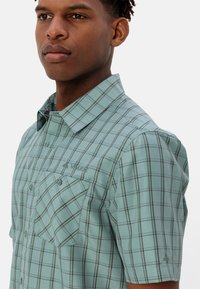 Korte mouwen geruit shirt in licht teal met donker groene en crème checks. Voorzien van twee borstzakken en het "Vaude" logo aan de voorkant.