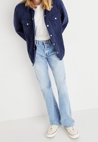 Giacca blu navy con bottoni sopra una t-shirt bianca, jeans a zampa d'elefante azzurri chiari e sneakers beige. Tessuto morbido con una leggera texture, vestibilità casual.