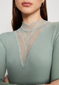 Halenka s dlouhým rukávem v měkké mátově zelené tkanině s vysokým výstřihem, která je zdobena jemným krajkovým detailem tvořícím tvar písmene V na přední straně.