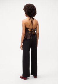 Haut halter en tissu texturé marron avec un design à peplum ; associé à un pantalon noir à jambes larges et des ballerines rouges. La vue arrière dévoile le détail du dos noué.