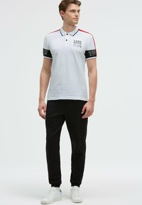 Biała koszulka polo z czerwonymi i czarnymi akcentami, posiada logo i numery na rękawach. Noszona z czarnymi dresami i białymi sneakersami.