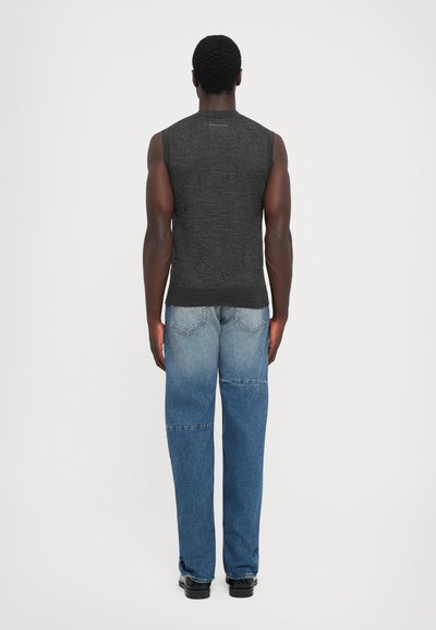 Uomo che indossa un canottiera grigia scura e jeans patchwork blu, in piedi di spalle contro uno sfondo chiaro e semplice.