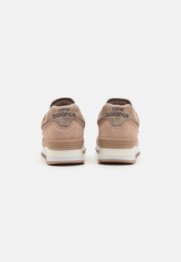 Ett par beige New Balance-sneakers visade bakifrån, i mocka med vit och brun sula mot en vit bakgrund.