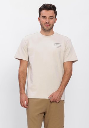 Man draagt een beige T-shirt met korte mouwen en ronde hals, met kleine geprinte tekst op de borst, en lichtbruine, gestructureerde broek tegen een effen achtergrond.