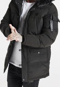 Svart puffjacka med foder i päls, med en dragkedjeficka på ärmen och knäppte framfickor. Strukturerad tyg och logotypdetalj.