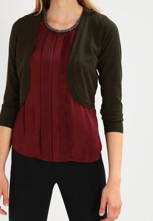 Vrouw in een donkergroene cropped cardigan over een geplooide bordeauxblauwe blouse, gecombineerd met zwarte broek, staand tegen een witte achtergrond.