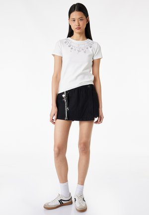 Jeune femme portant un t-shirt blanc à manches courtes avec des motifs d'étoiles, une mini-jupe noire avec un détail chaîne, des chaussettes blanches et des baskets blanches.