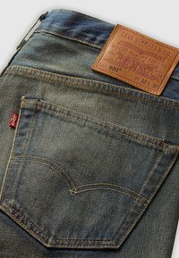 Blue denim jeans feature a leather patch with brand logo and size, contrasting orange stitching, and a distinctive back pocket design.