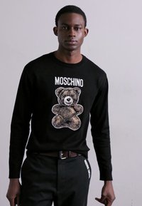 Fekete pulóver, amelyen egy gráfika látható egy maci képpel, és fehér betűkkel a "MOSCHINO" felirattal a minta felett. A modellen szabott fekete nadrág van.