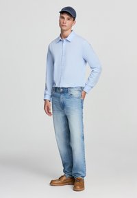 Camicia button-up azzurro chiaro, jeans a lavaggio chiaro, scarpe casual marroni e un cappellino blu. Design semplice con vestibilità dritta e senza modelli visibili.