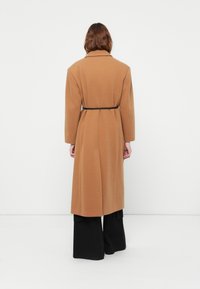 Caban lung de culoare cămilă cu un design structurat, având talie cu curea, mâneci largi și un material texturat, asortat cu pantaloni largi negri.